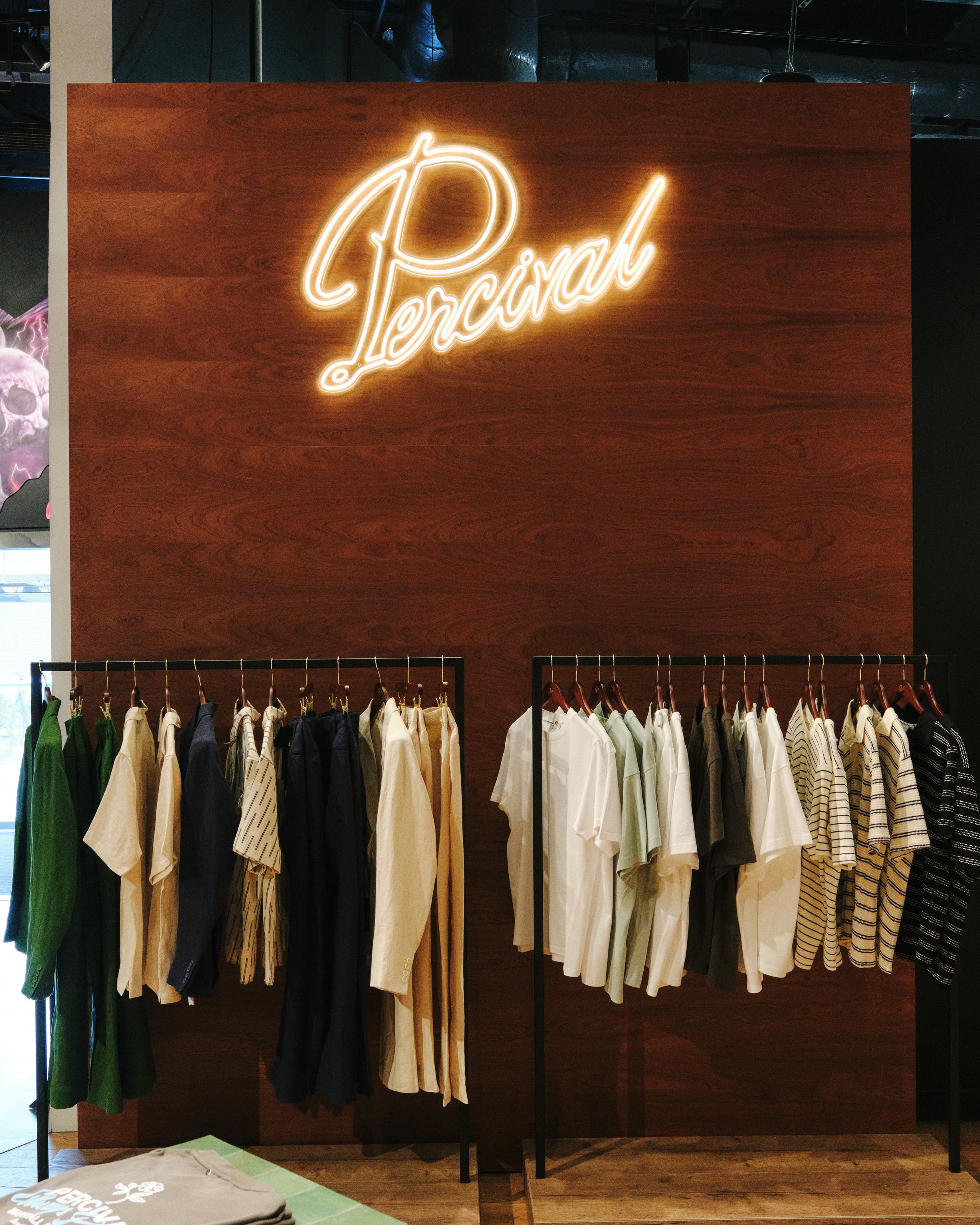 Glowing Percival neon brand sign on a wooden wall panel above rows of men's clothing racks at the Selfridges Birmingham pop-up store.