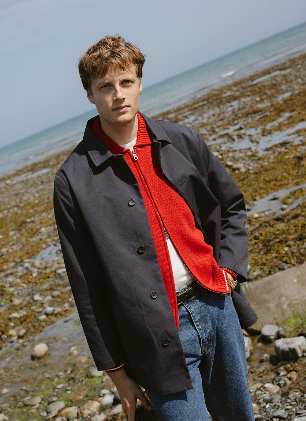 A model wearing a Percival trench coat over a red zip cardigan and blue jeans by the coast.