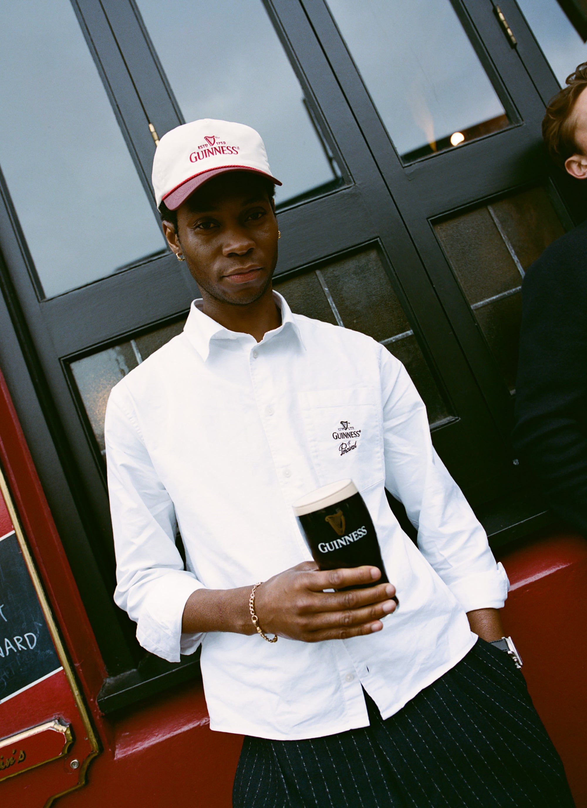 A man models the white Guinness x Percival button-down shirt, holding a pint of Guinness outside a pub.