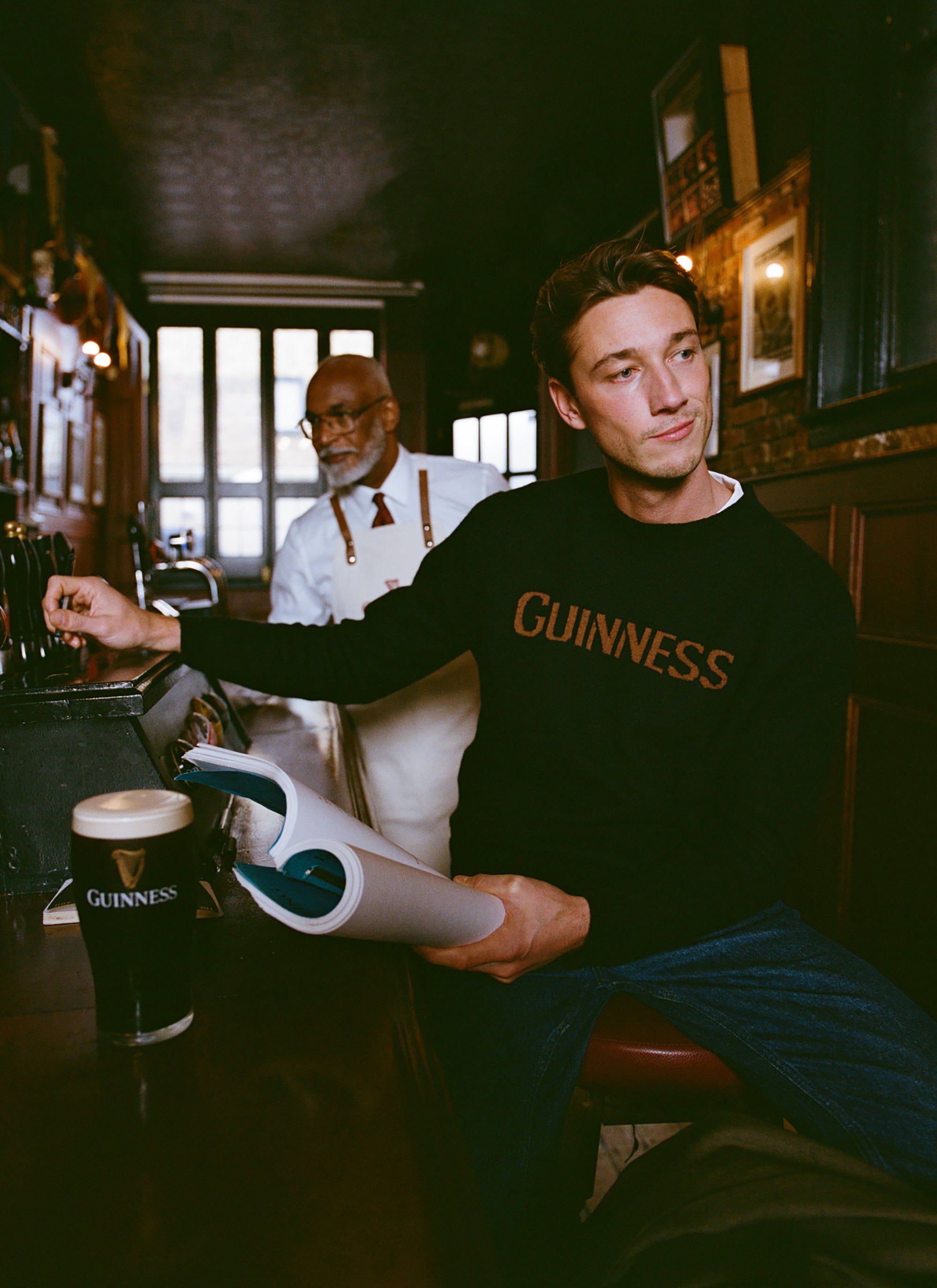 A man wearing the navy Guinness x Percival jacquard jumper sits at a pub bar, with a pint of Guinness and a bartender nearby.
