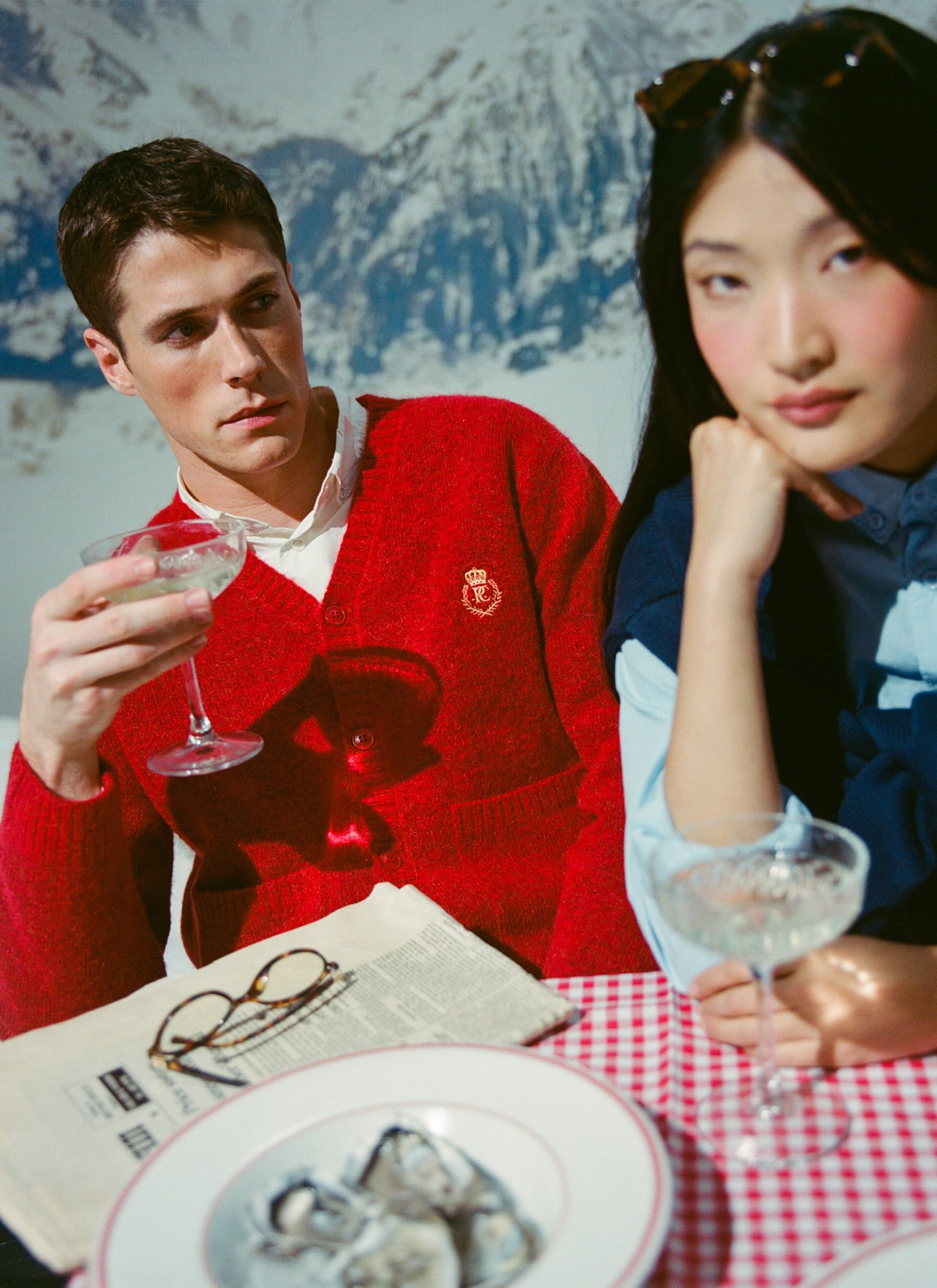 A man models the red Champion x Percival mohair cardigan, enjoying champagne with a companion in an après-ski setting.