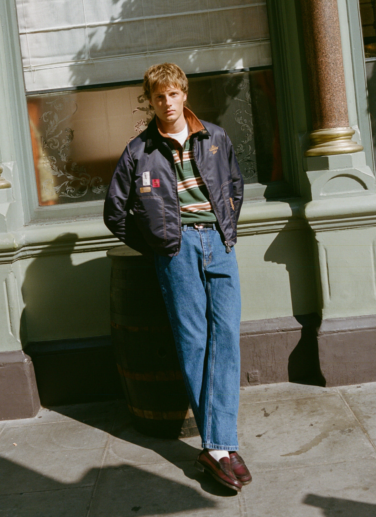 Model wearing the Guinness x Percival navy bomber jacket, striped rugby shirt, and blue jeans leaning against a pub barrel.