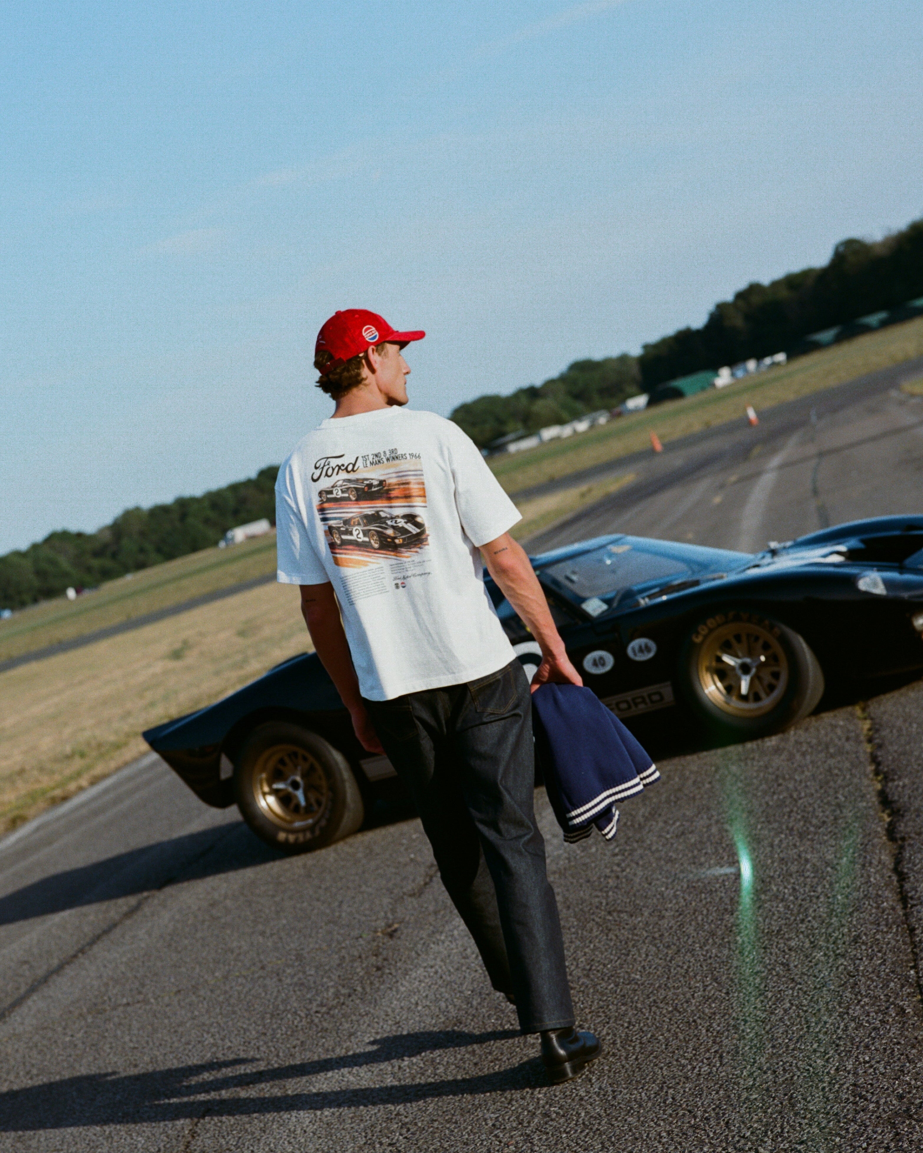 A man wearing a white Ford x Percival t-shirt, dark pants, and a red cap walks on an airfield road next to a black classic Ford GT car, holding a blue jacket.