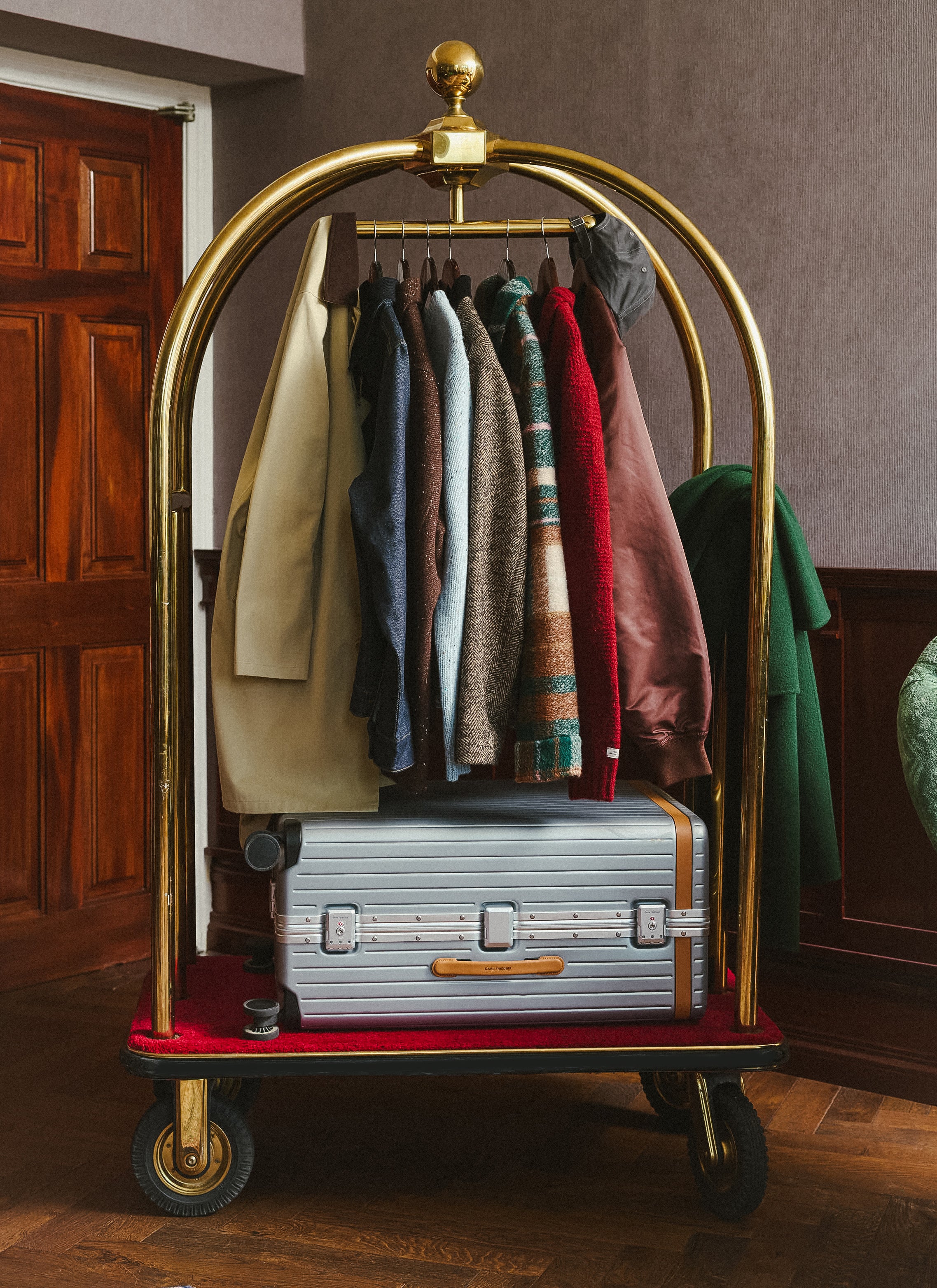 A gold luggage trolley featuring various Percival AW26 coats, jumpers and outerwear hanging above a silver suitcase.