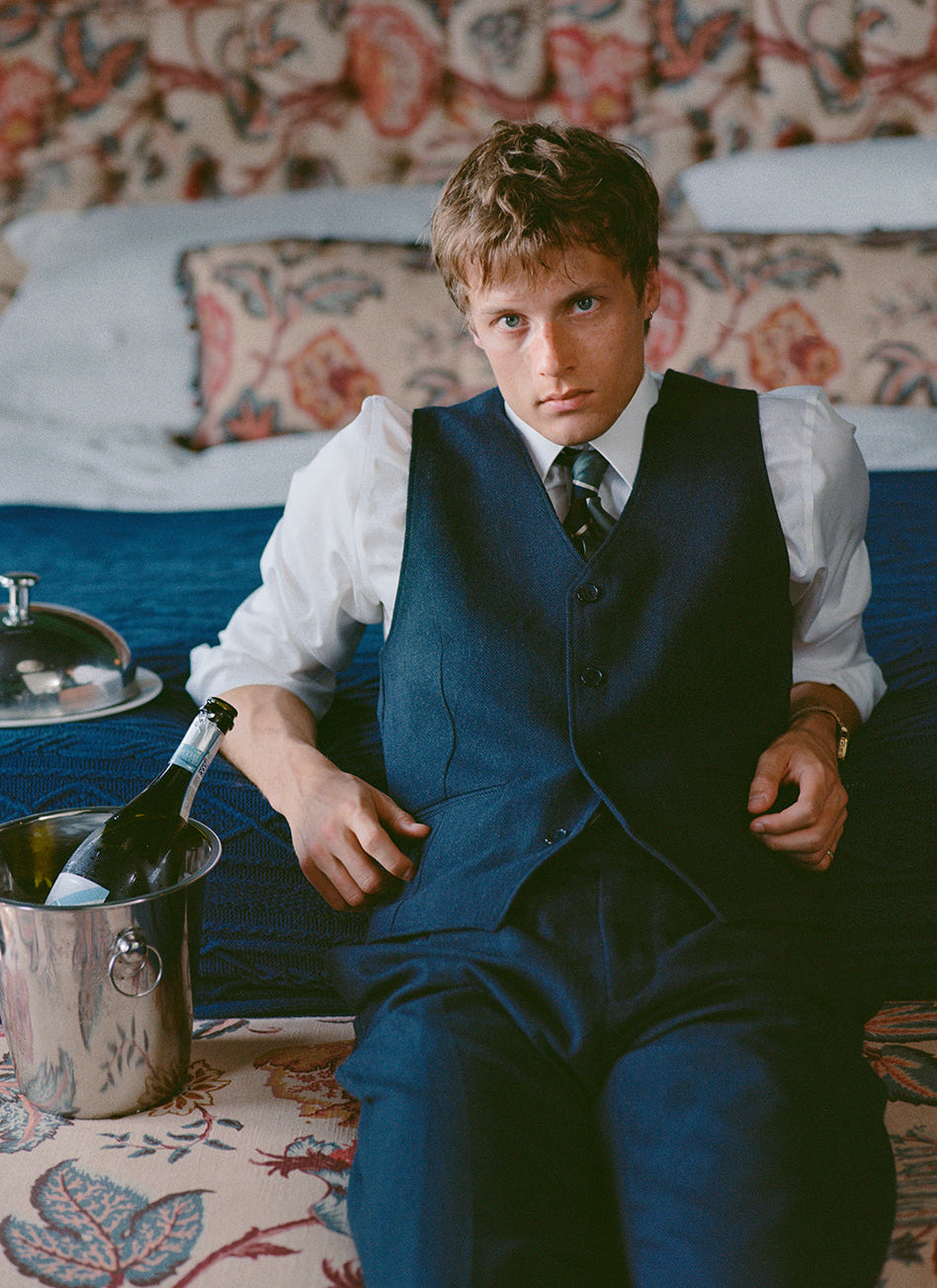 A model wears the Percival Waistcoat and Percival Stanley Pleated Tailored Trousers in Navy Herringbone Wool in a hotel room.