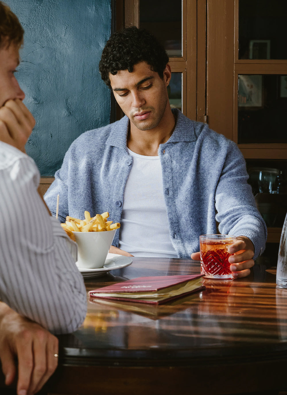 A man sits at a table wearing the heather blue Percival Archive Knit Collared Cardigan open over a white vest.