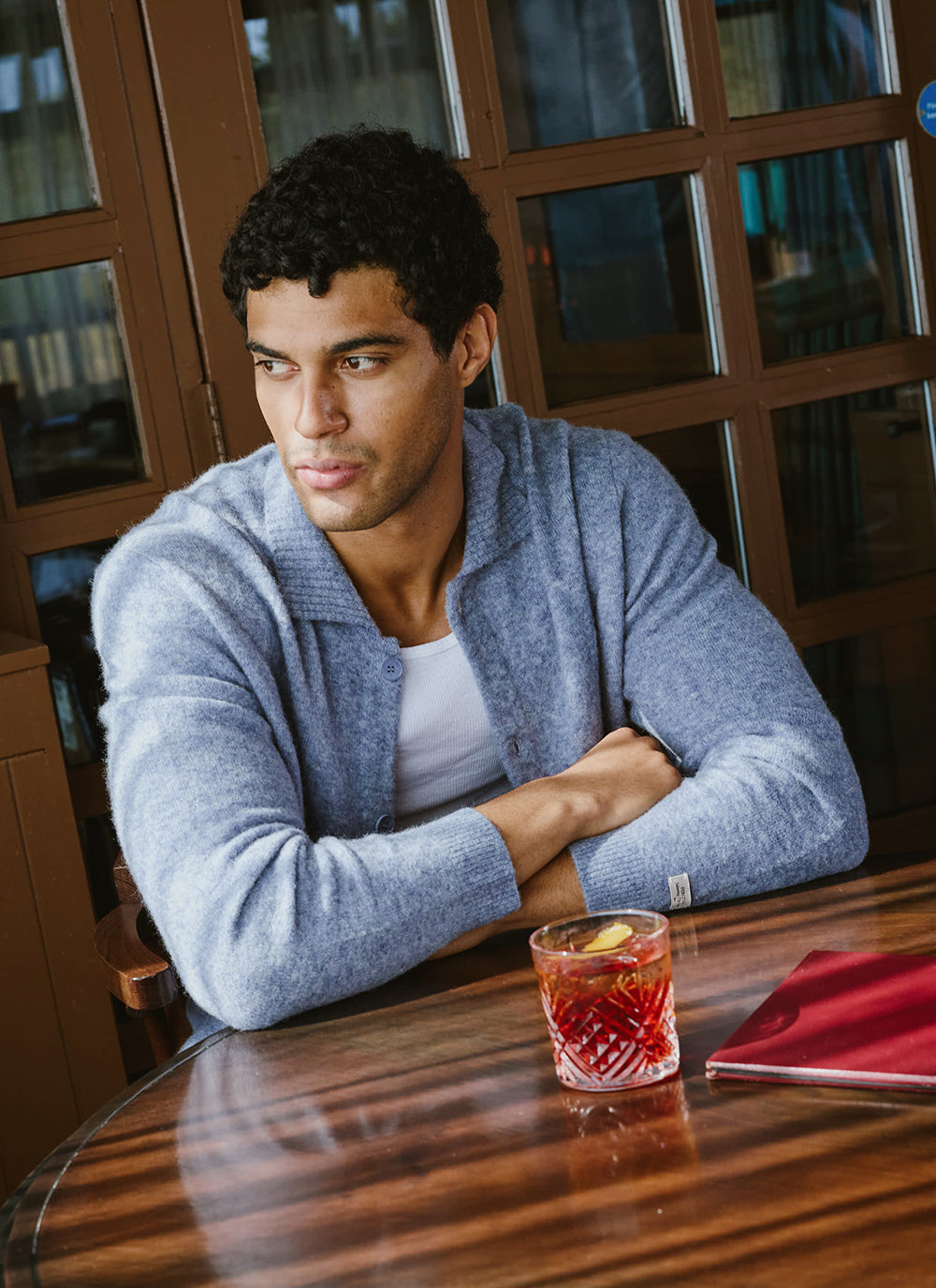 A model wears the heather blue Archive Knit Collared Cardigan from Percival, seated at a table with a cocktail.