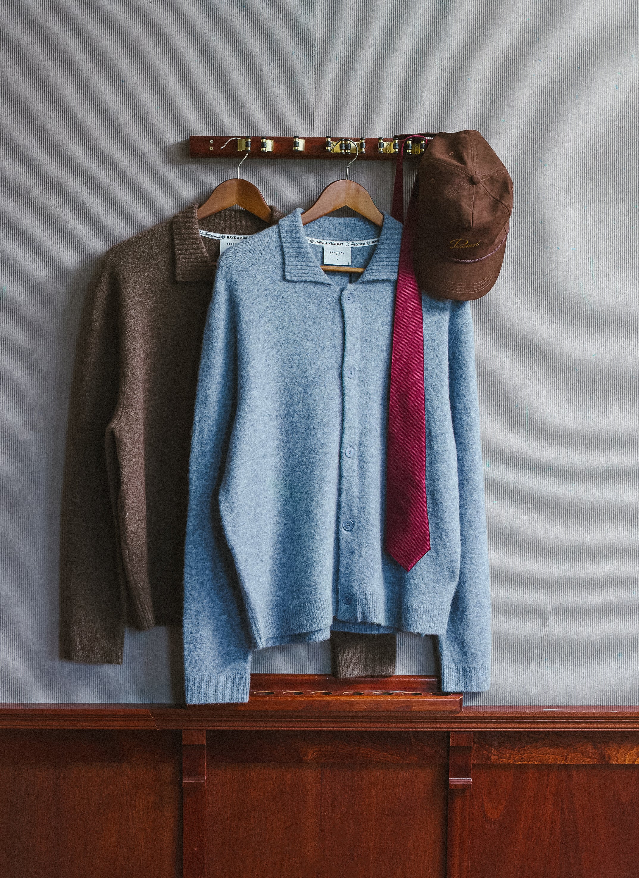 Percival archive knit collared cardigans in brown and heather blue, hanging alongside a brown trucker cap.