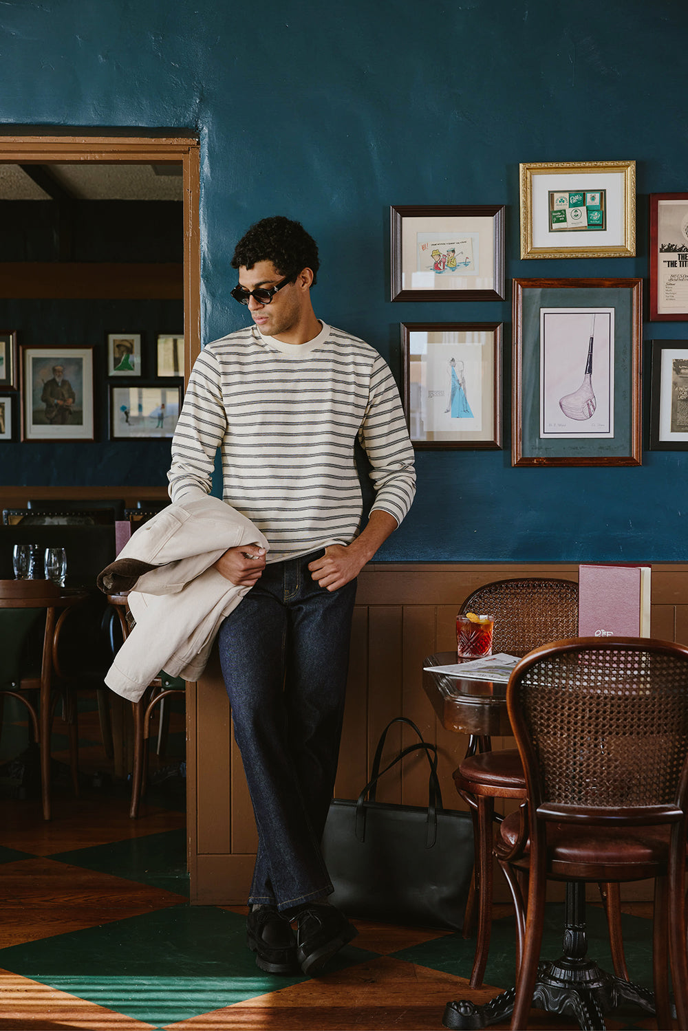 A model wears the white Percival Arlo Breton long-sleeve t-shirt, holding a jacket inside a pub with blue walls.