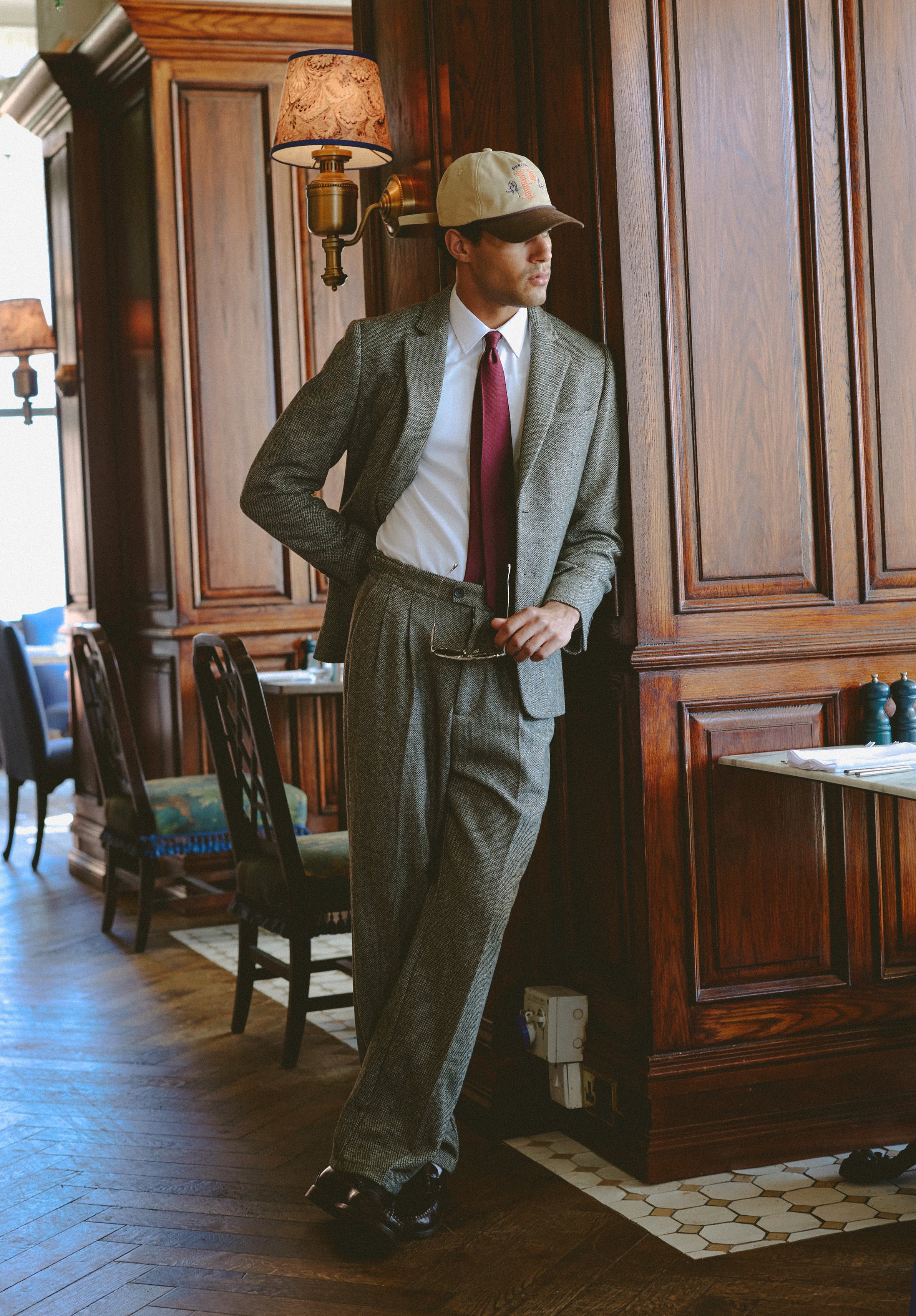 A model wears the Percival grey birdseye wool suit, styled with a burgundy tie and cap in a wood-panelled interior.