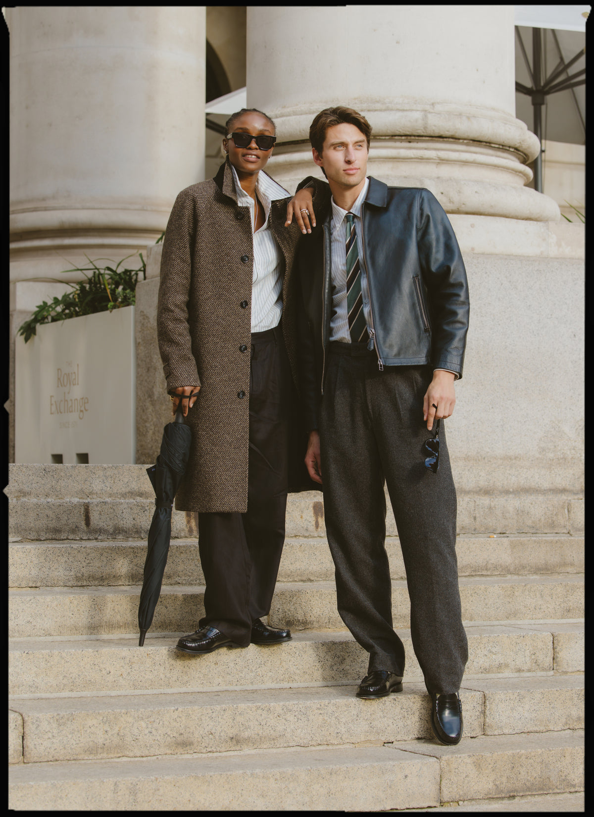 Two models standing on outdoor stone steps, wearing Percival charcoal tailored trousers, a car coat, and a leather jacket.