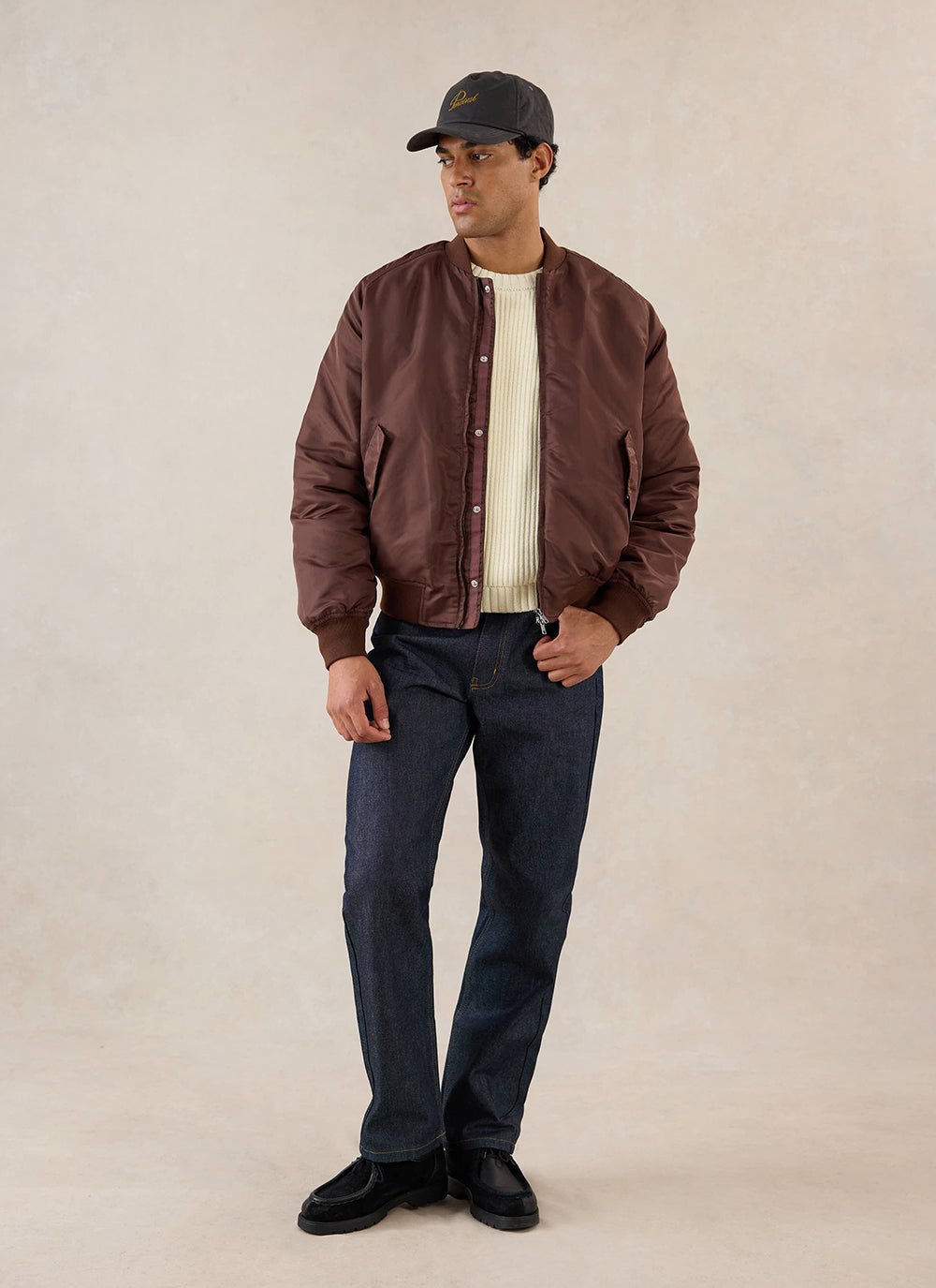 A model wears the espresso Percival nylon bomber jacket over a cream knit jumper and dark trousers in a studio.
