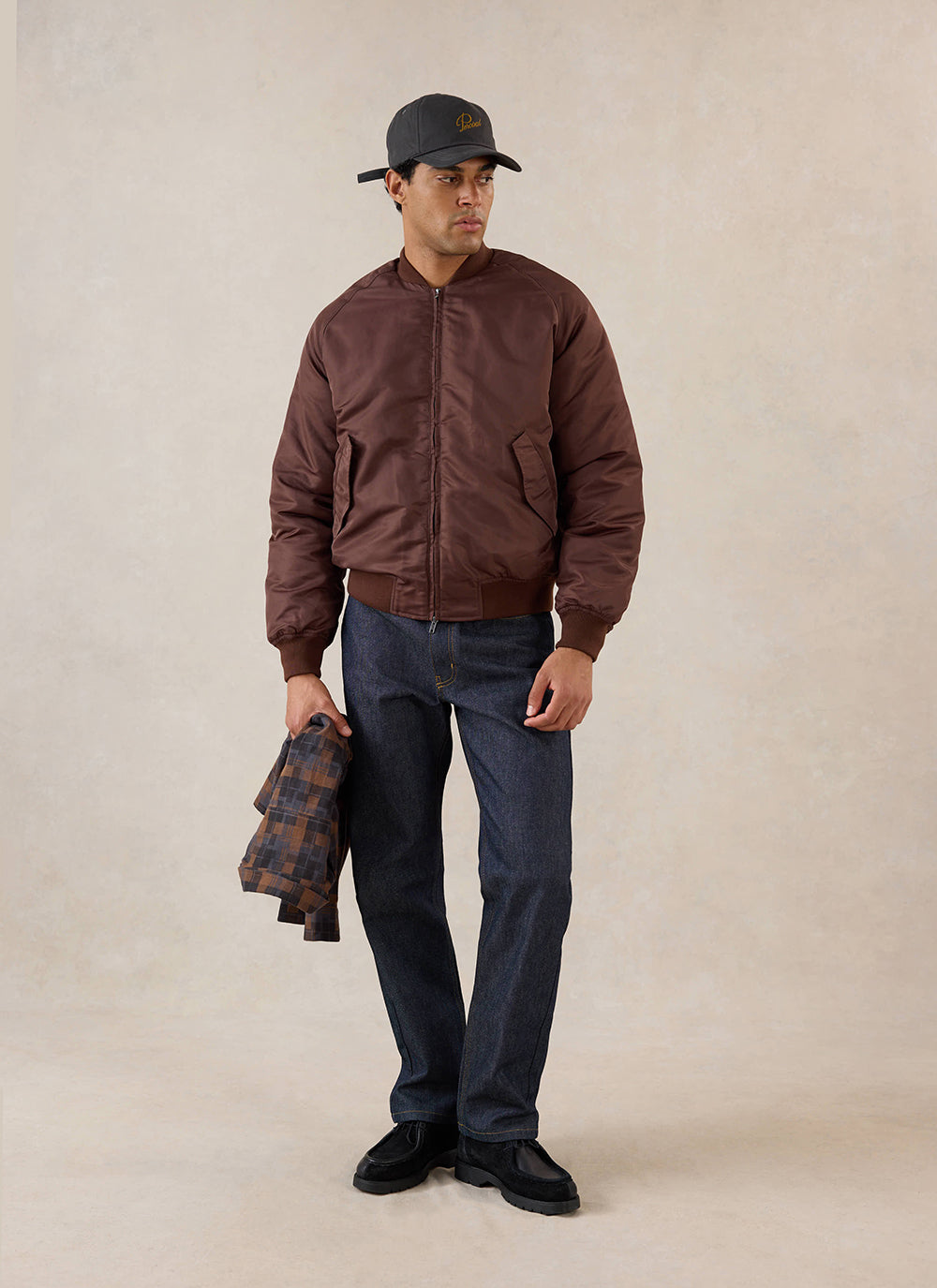 A model wears the espresso nylon bomber jacket from Percival over dark denim jeans and a cap, holding a patterned shirt.