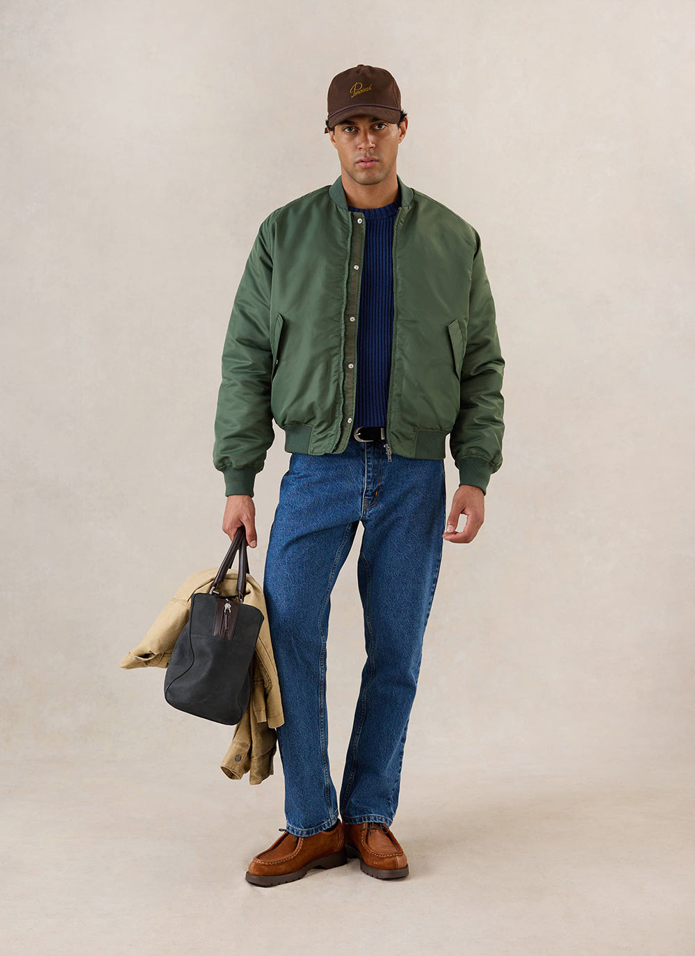A model wears the forest nylon bomber jacket from Percival over a blue knit jumper and jeans in a studio shot.