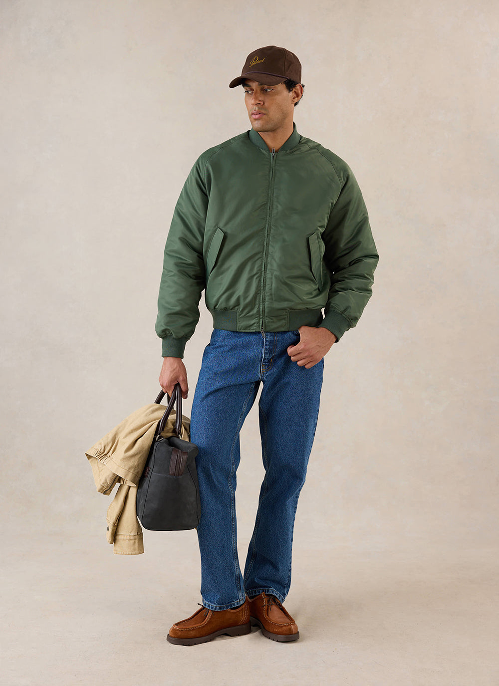 A model wears the forest green nylon bomber jacket from Percival with blue jeans, a cap, and a bag in a studio.