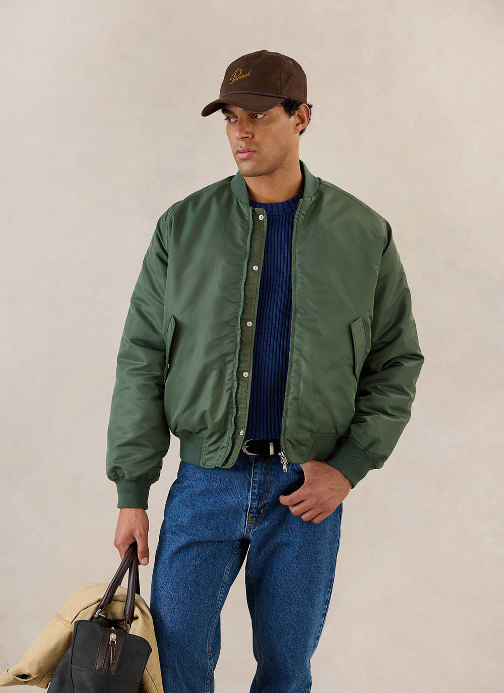 A model wears the forest green Percival nylon bomber jacket over a blue knit jumper and blue jeans, holding a bag.