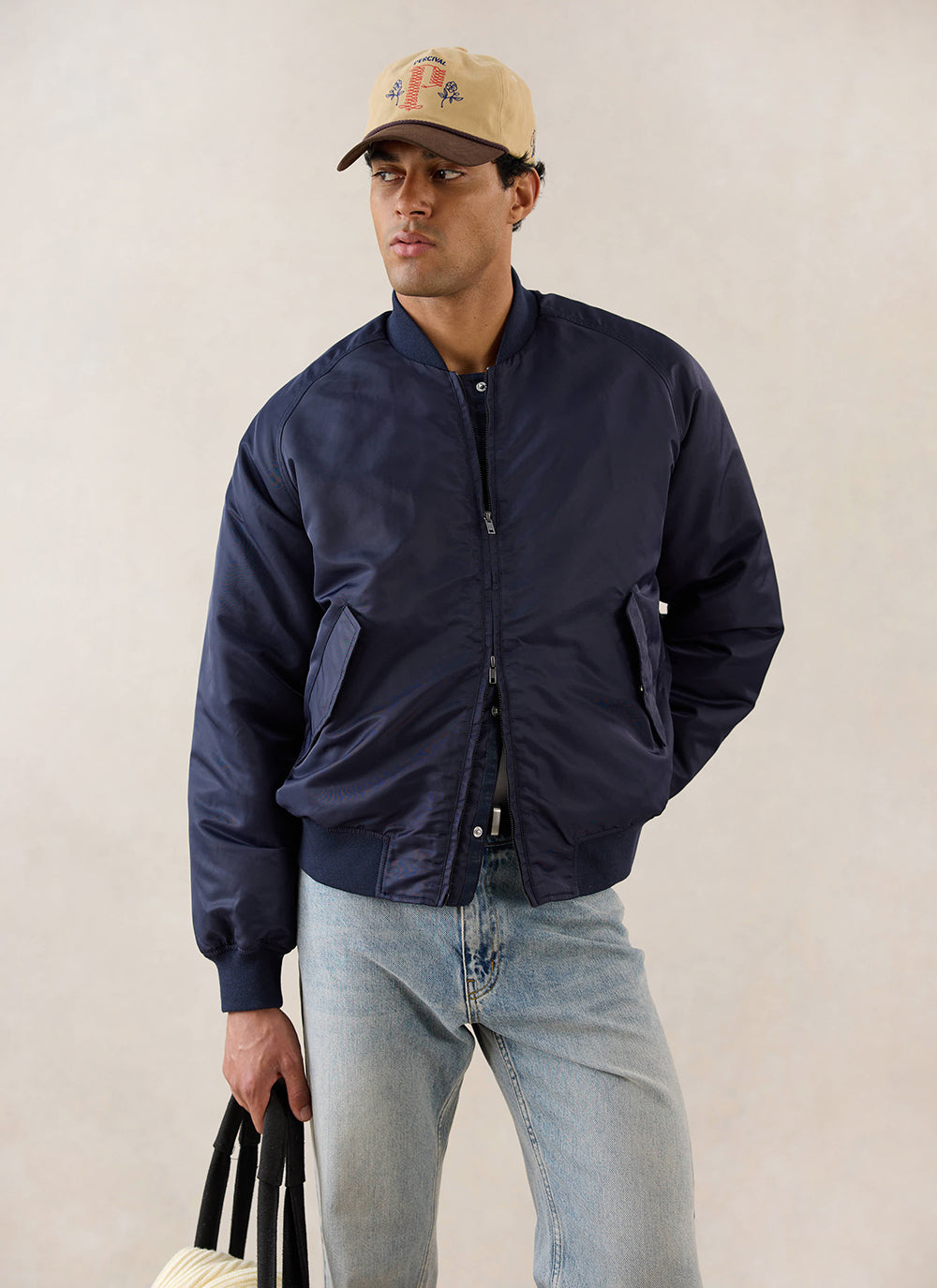 A model wears the midnight blue Percival nylon bomber jacket with light wash jeans and a cap in a studio.