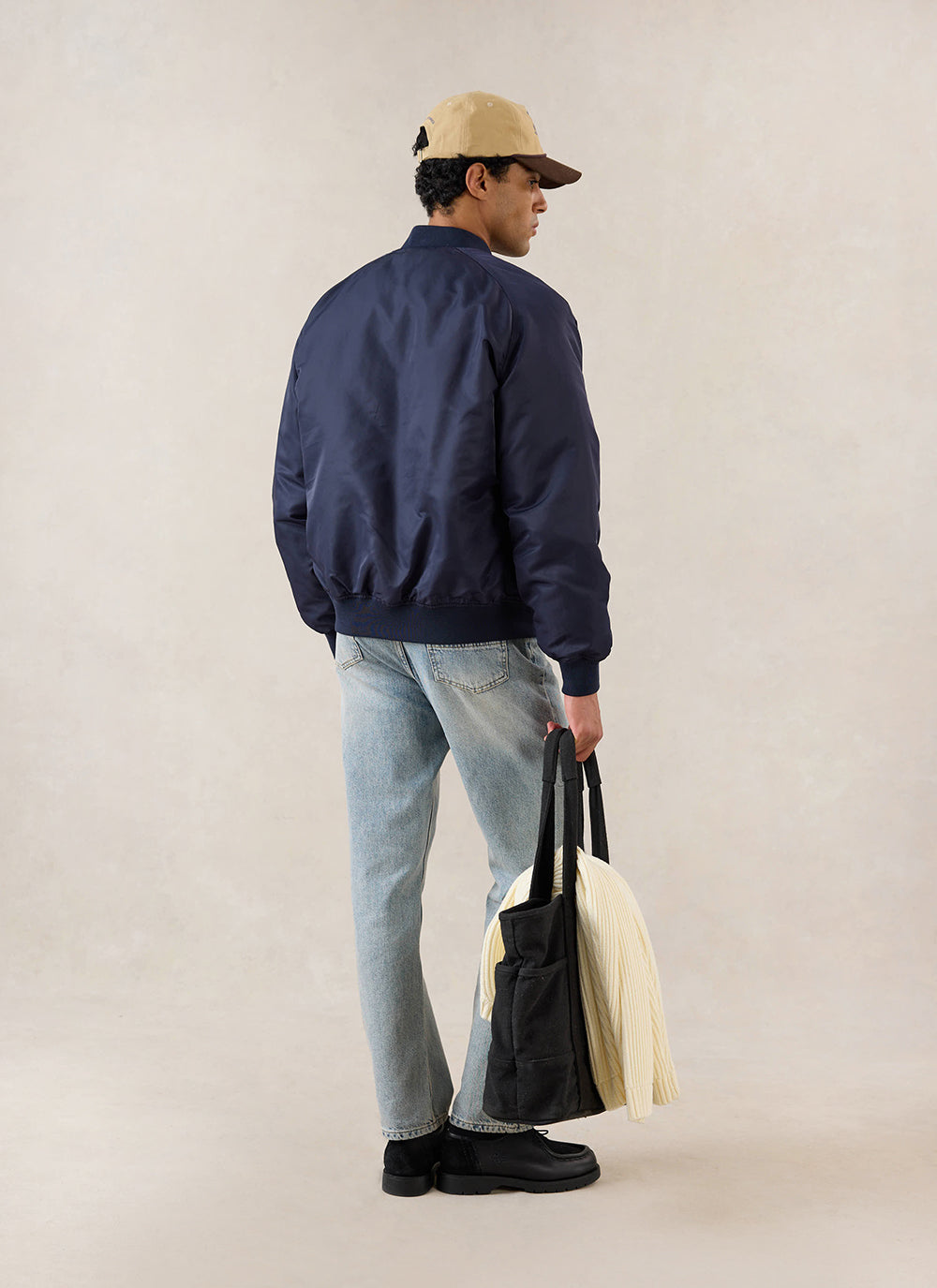 A model from behind wearing the midnight blue Percival nylon bomber jacket with light jeans and a tote bag in a studio.