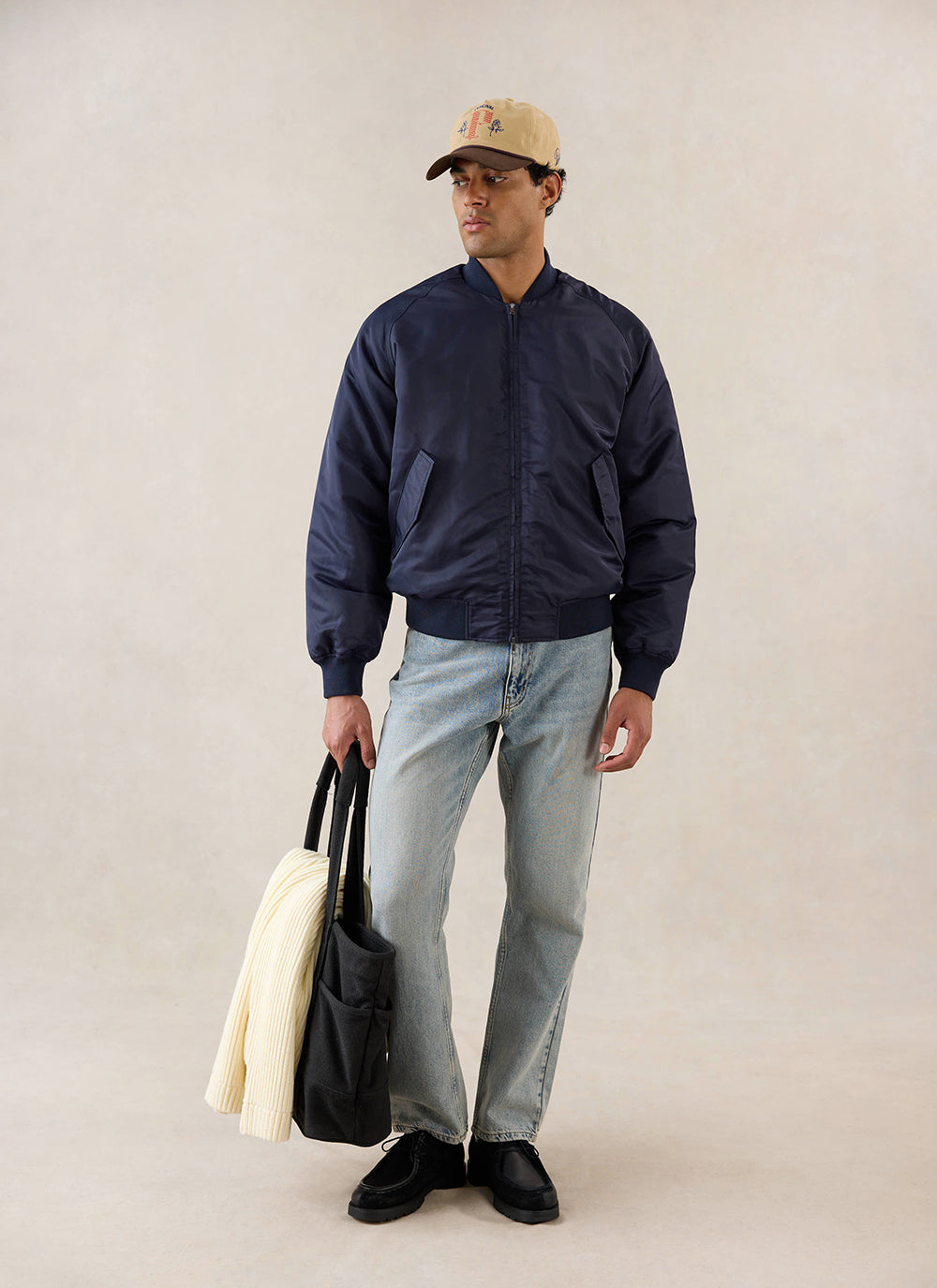 A model wears the midnight nylon bomber jacket from Percival over light jeans, holding a black bag and cap in a studio.