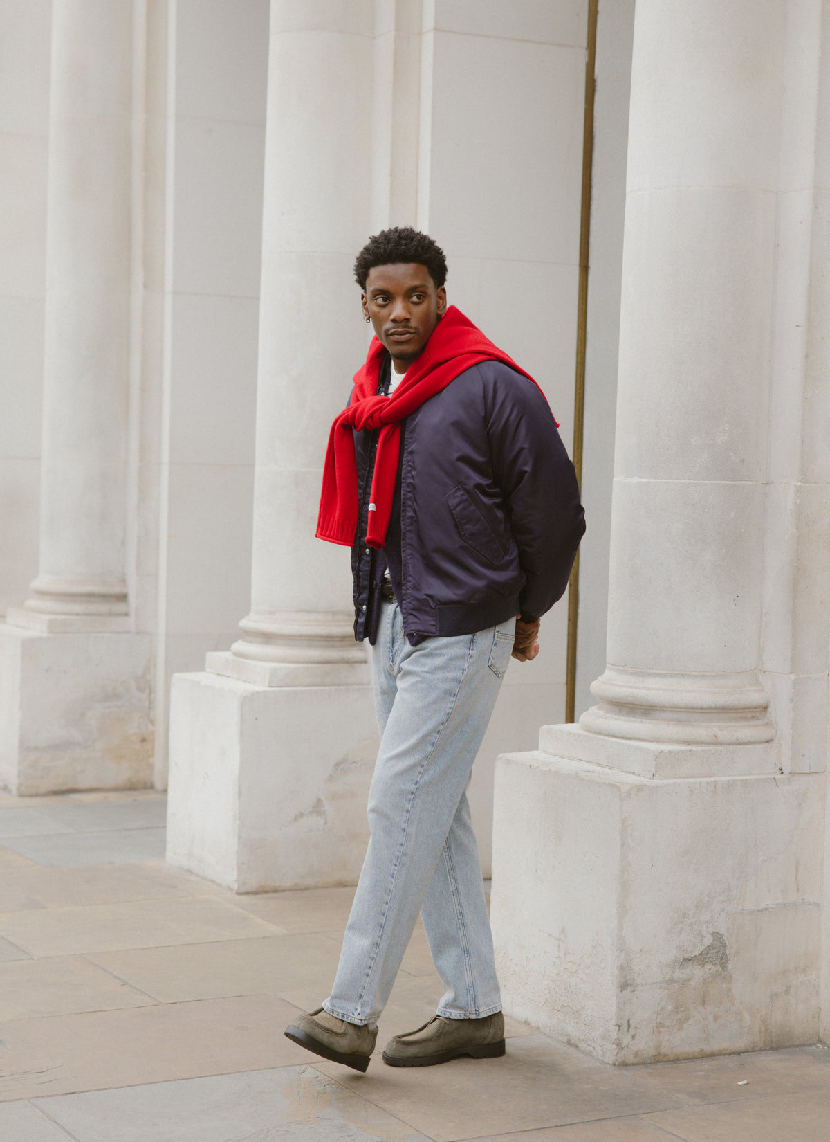 A model wearing a dark bomber jacket with the red jumper draped over his shoulders outdoors near large stone pillars.