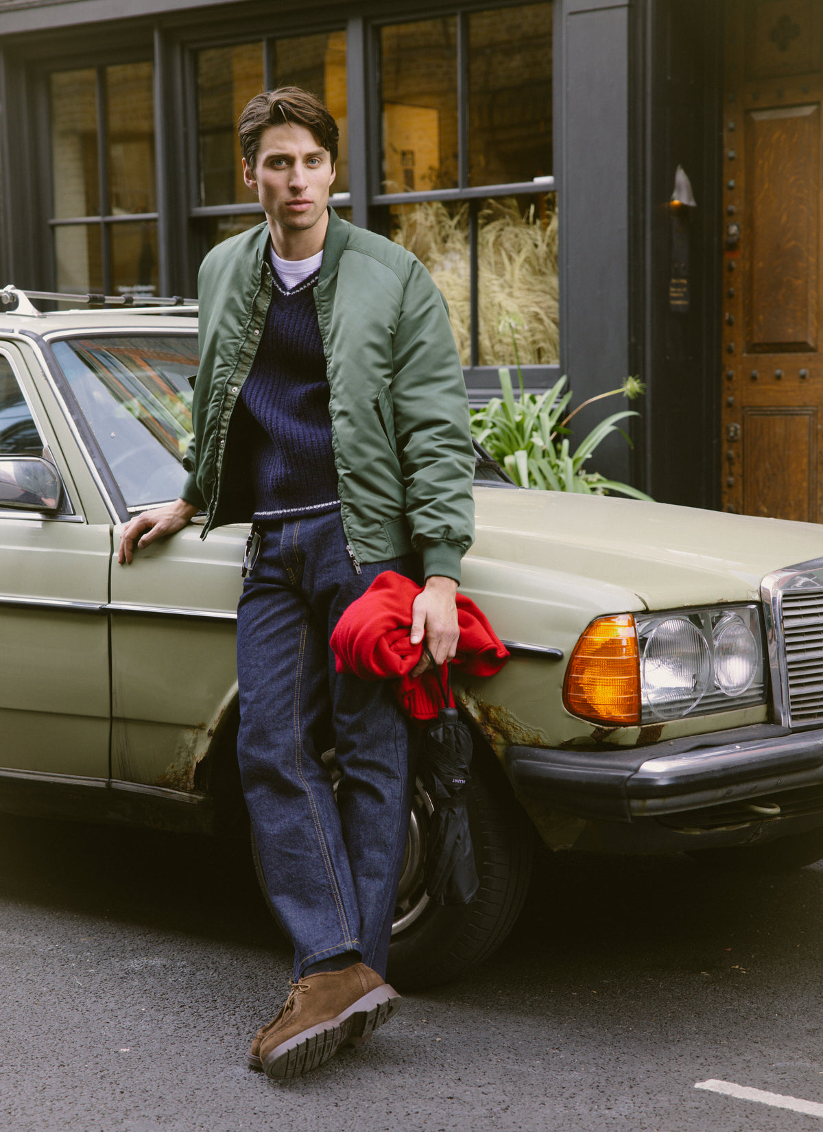 A model leaning against a vintage car wearing the Percival navy V-neck jumper under a green bomber jacket.