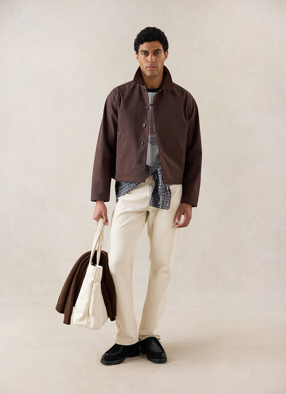A model wears the espresso Percival Carter Cropped Trench Coat with cream trousers and a tote bag in a studio.