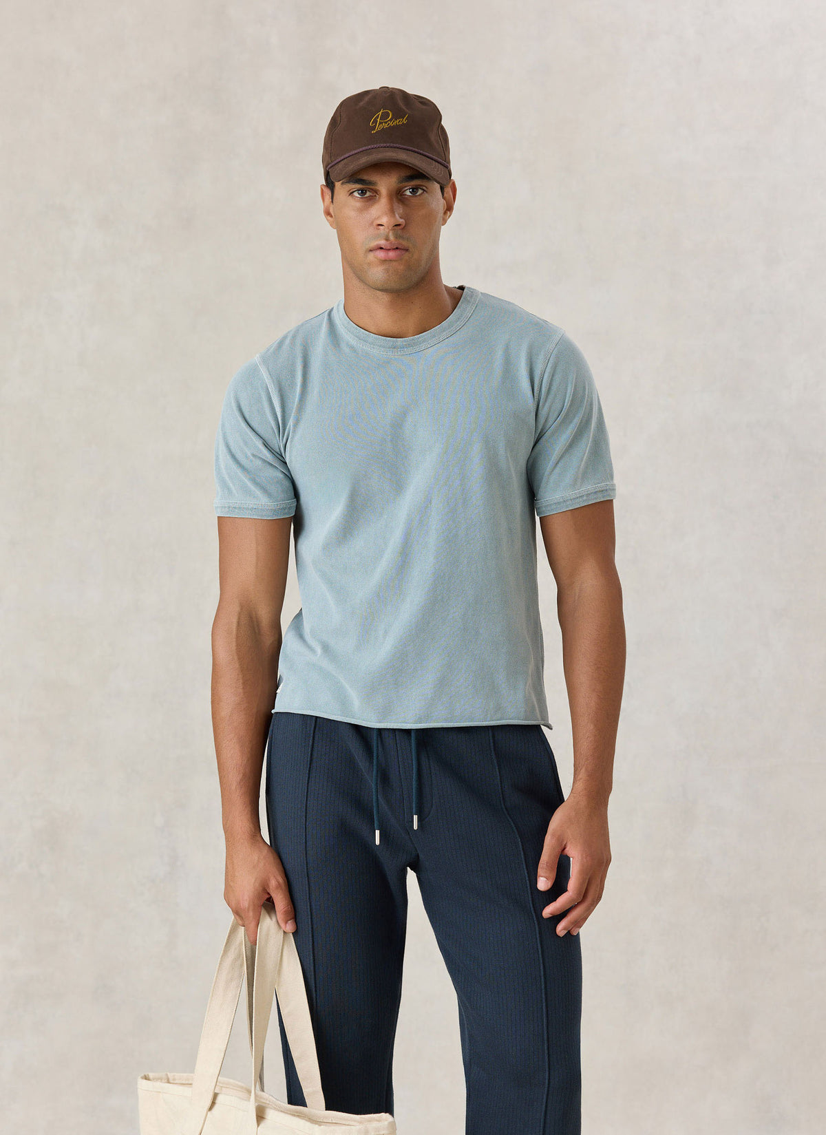 A model wearing the light blue Percival Casey vintage wash cropped t-shirt with dark trousers, a cap, and a tote bag.