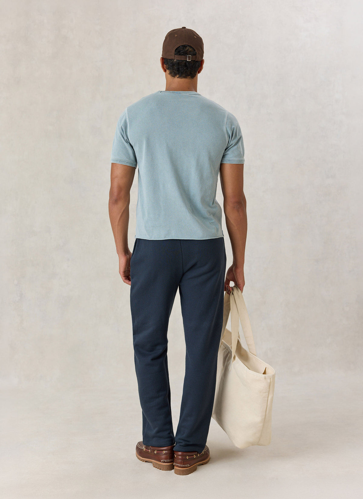 A model from the back wearing the Percival light blue vintage wash cropped t-shirt with dark trousers and a canvas tote bag.