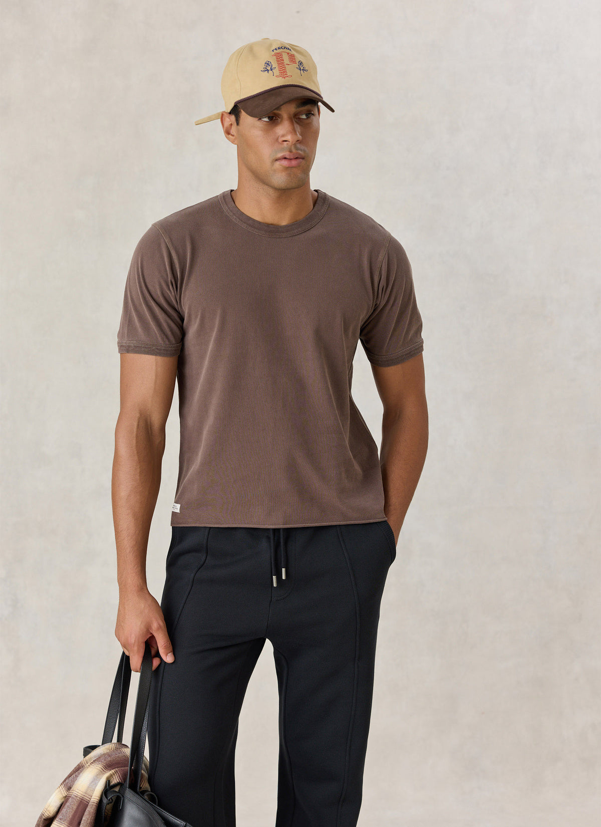 A model wearing the brown Percival Casey vintage wash cropped T-shirt with a cap, dark trousers, and a bag indoors.