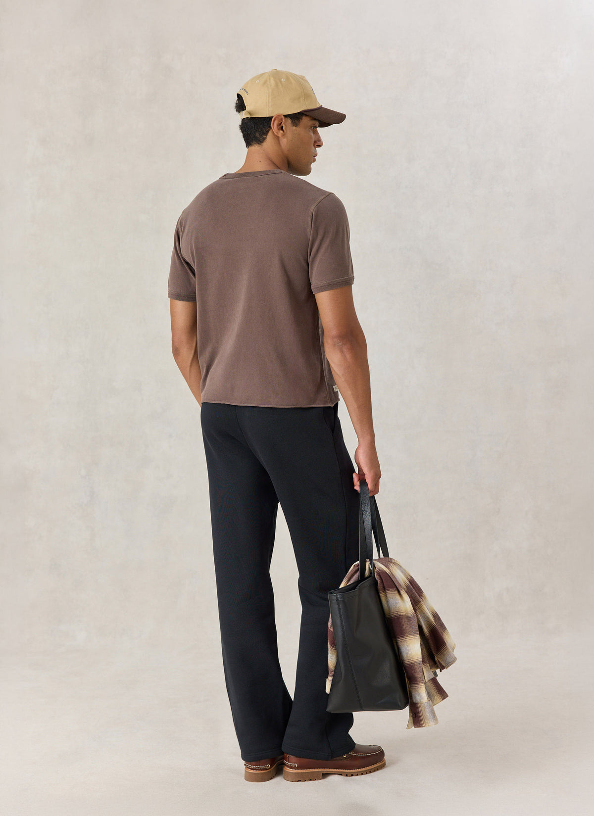 A model from the back wearing the Percival brown vintage wash cropped T-shirt, holding a black tote bag and plaid shirt.