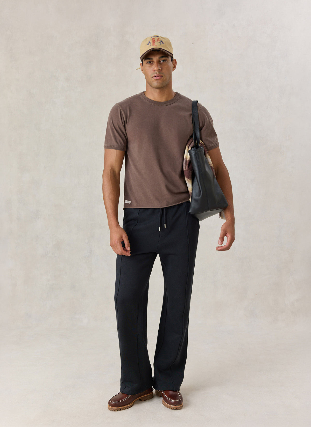 A model wearing the brown Percival Casey vintage wash cropped t-shirt with black trousers, a cap, and a leather tote bag.