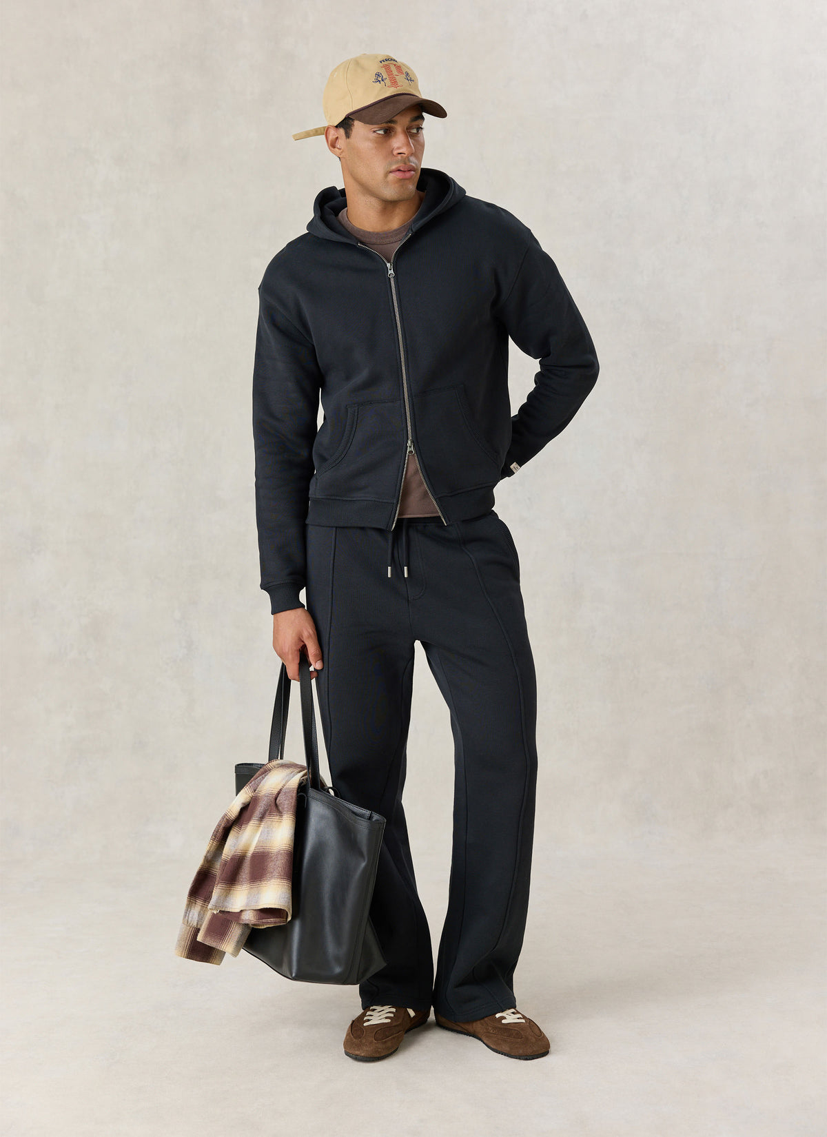 A model wearing Percival black cotton zip hoodie and matching trackpants, with a cap and a black tote bag.