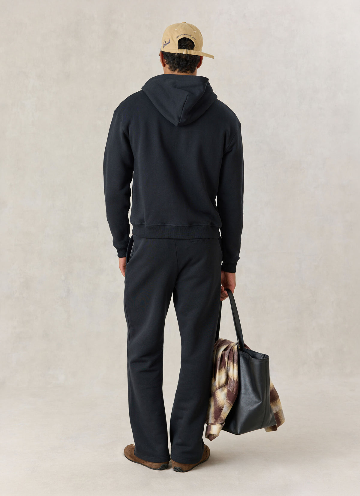 A model from behind wearing a black Percival zip hoodie and trackpants, holding a tote bag with a plaid shirt.