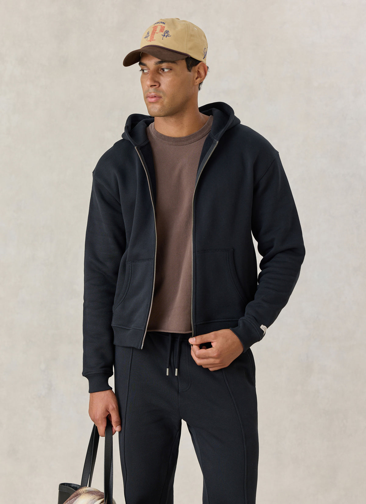 Model wearing the Percival black cotton jersey zip hoodie with matching trackpants, a brown t-shirt, and a cap.