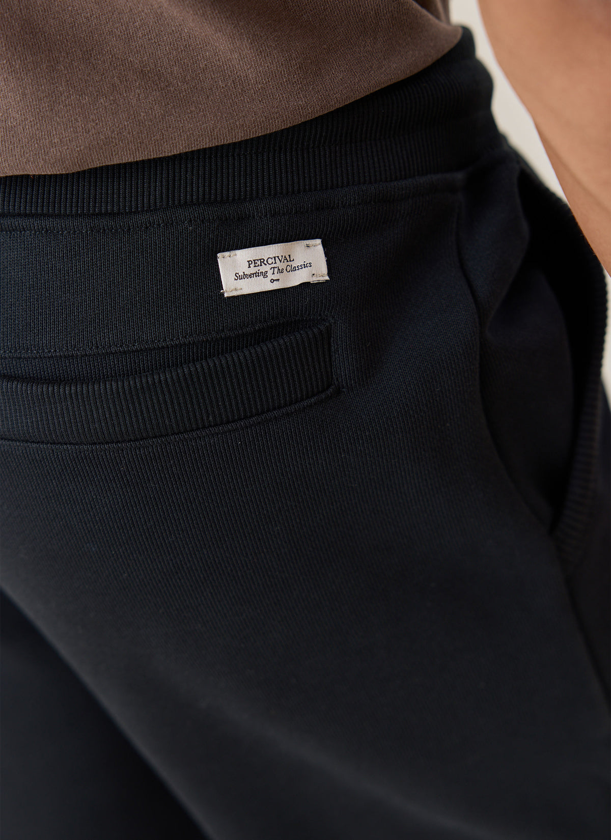A close-up of the Percival black organic cotton jersey trackpants showing the back pocket and woven brand label.