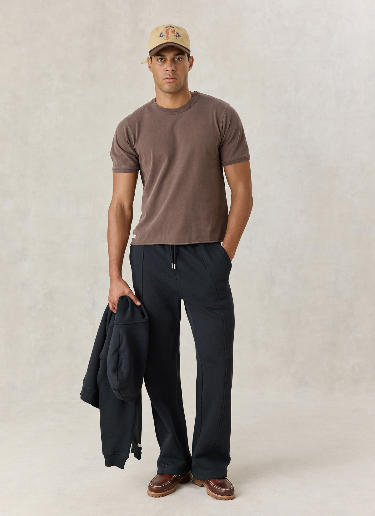A model wearing Percival black cotton jersey straight leg trackpants with stitched front creases, a brown t-shirt, and a cap.