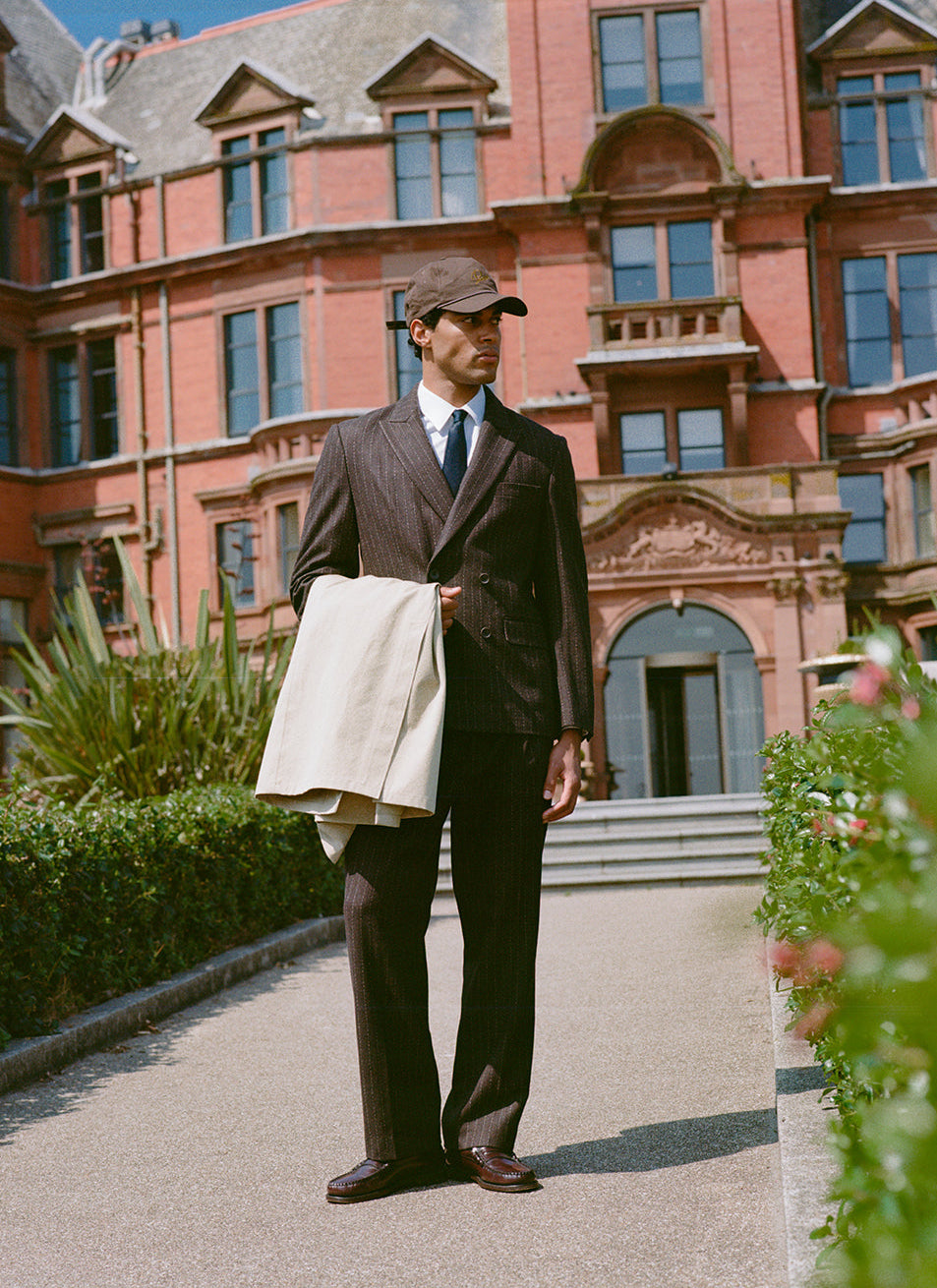 Model wears the Percival Clifton pinstripe suit and Luther trucker cap in espresso, standing outside a brick building.