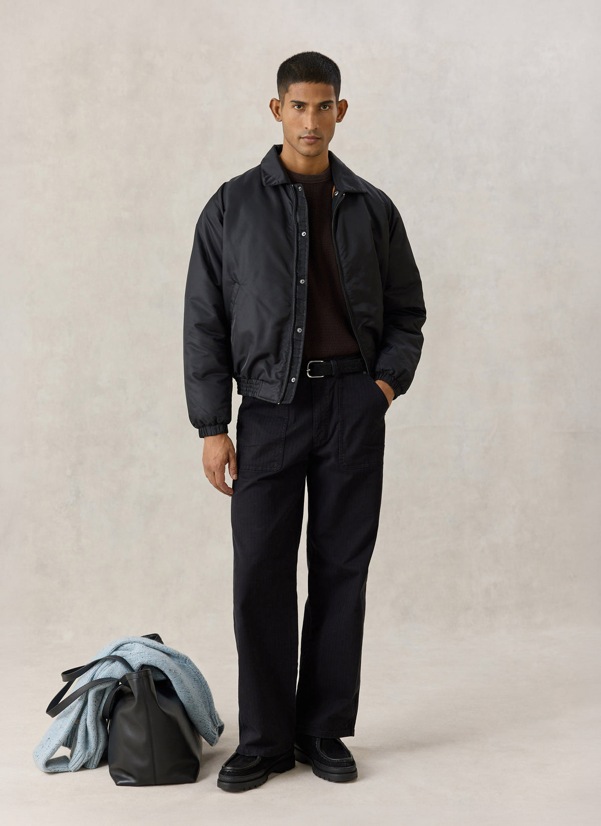 Model wearing the Percival black nylon collared bomber jacket with relaxed trousers, a dark knit, and a bag on the floor.