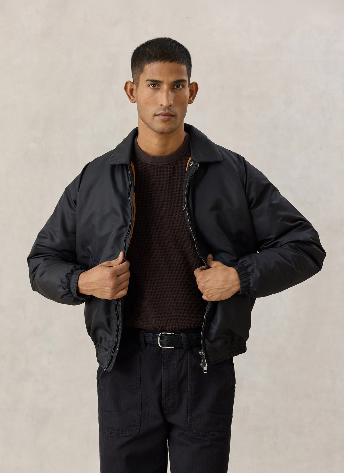 A model wearing the Percival black nylon collared bomber jacket with a quilted lining over a brown knit jumper.