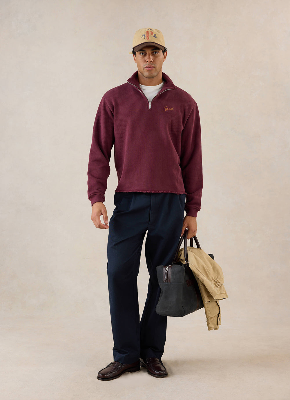 A model wears the burgundy Percival Conrad 1/4 zip jumper with dark trousers and a cap in a studio.