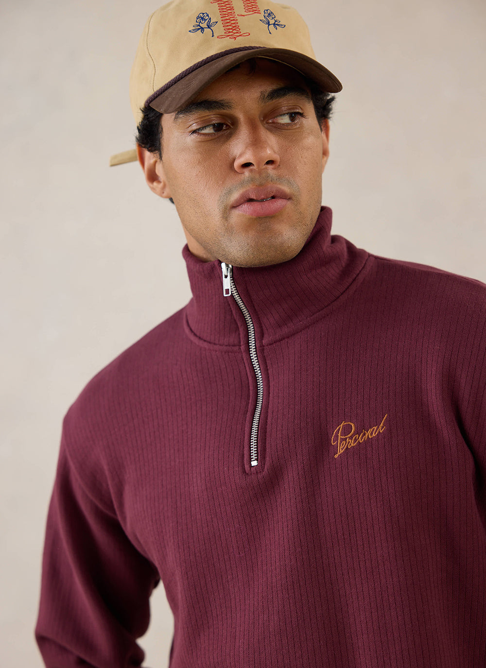 A close-up of a model wearing the burgundy Percival Conrad Funnel Neck 1/4 Zip Jumper and a cap, showing the collar detail.