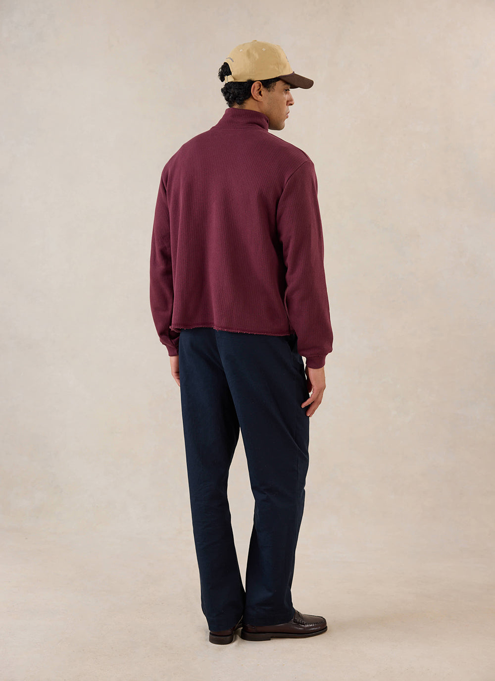 Rear view of a model in the burgundy Conrad 1/4 zip jumper from Percival, styled with dark trousers and a cap.
