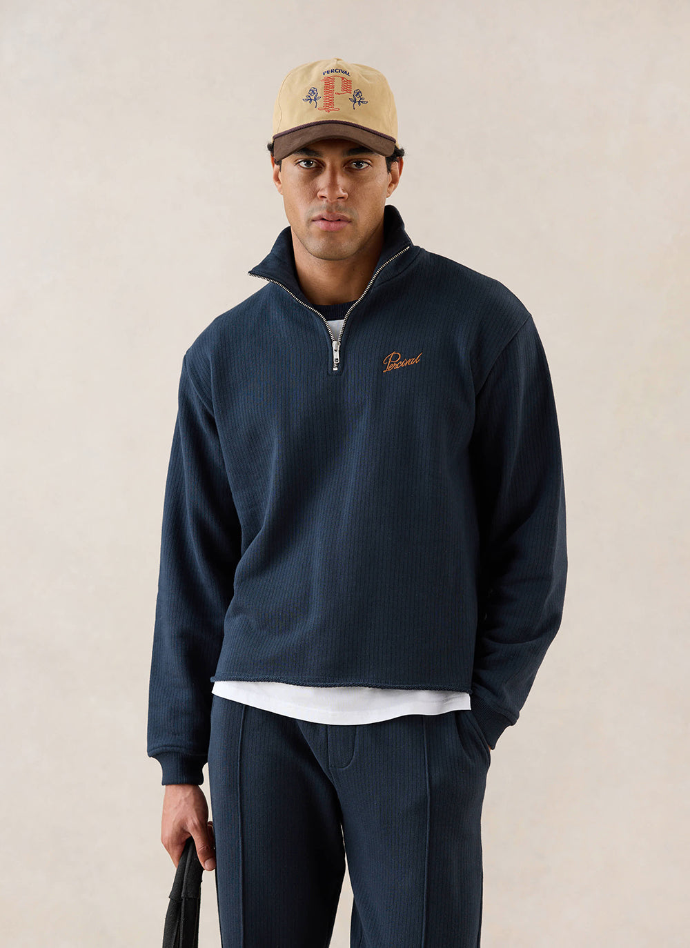 A model wears the navy Percival Conrad 1/4 zip funnel neck jumper, styled with matching trousers and a cap in a studio.