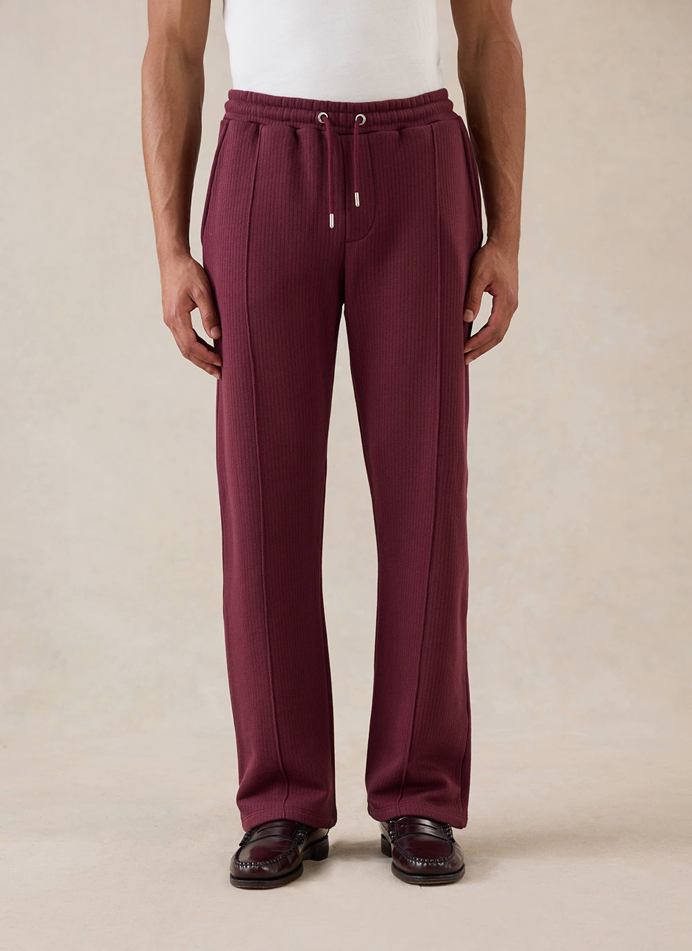 A model wears the merlot Percival Conrad straight leg trackpants, styled with a white t-shirt and dark loafers.