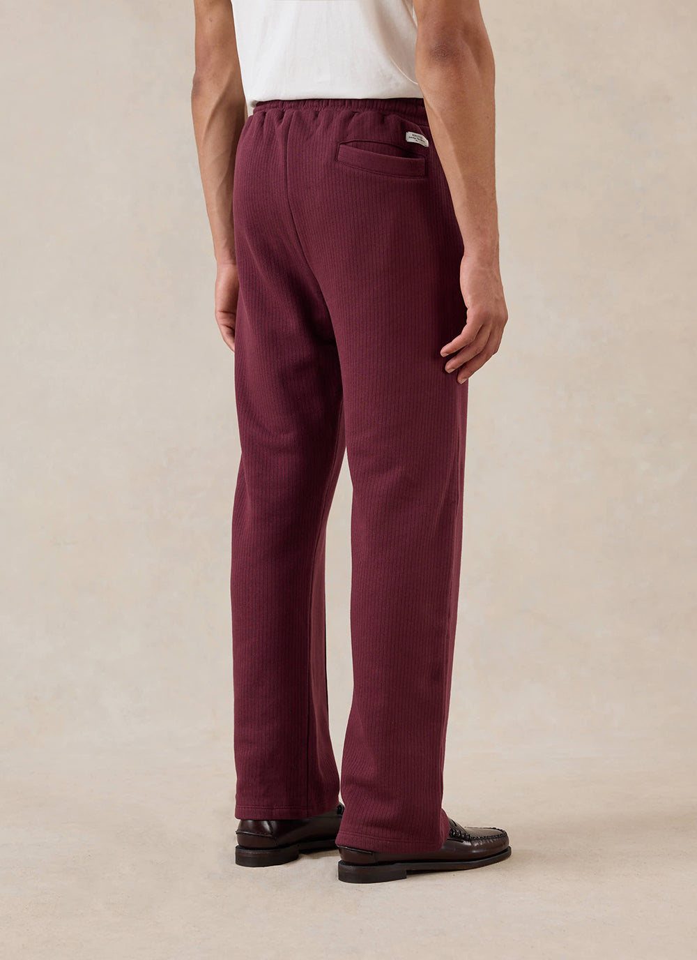 A model wears the merlot Percival Conrad straight leg trackpants from behind, paired with a white t-shirt and loafers.