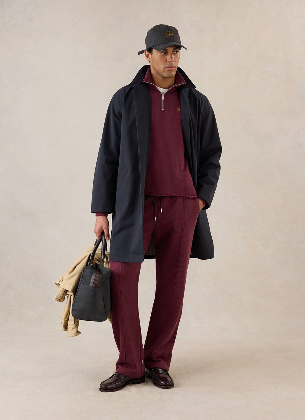 A model wears the merlot Percival Conrad trackpants, styled with matching jumper, dark coat, cap, and holdall.