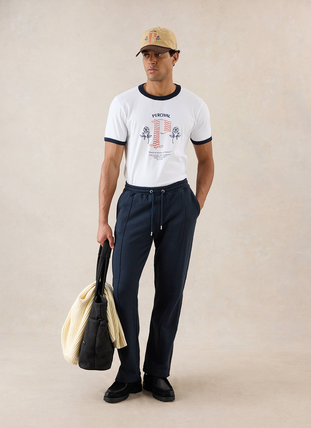 A model wears navy Percival Conrad trackpants, styled with a white ringer T-shirt, cap, tote bag and black shoes.
