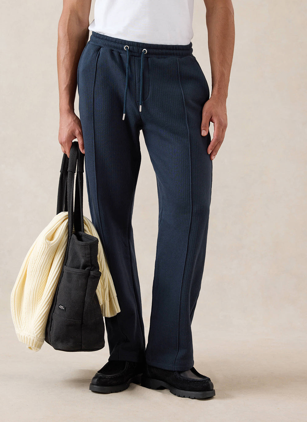 A model wears navy Percival straight-leg trackpants with a white t-shirt, holding a black tote bag in a studio shot.