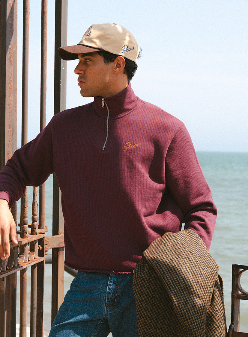 A model wears the burgundy Percival Conrad funnel neck 1/4 zip jumper in 100% cotton with a cap and a patterned coat.