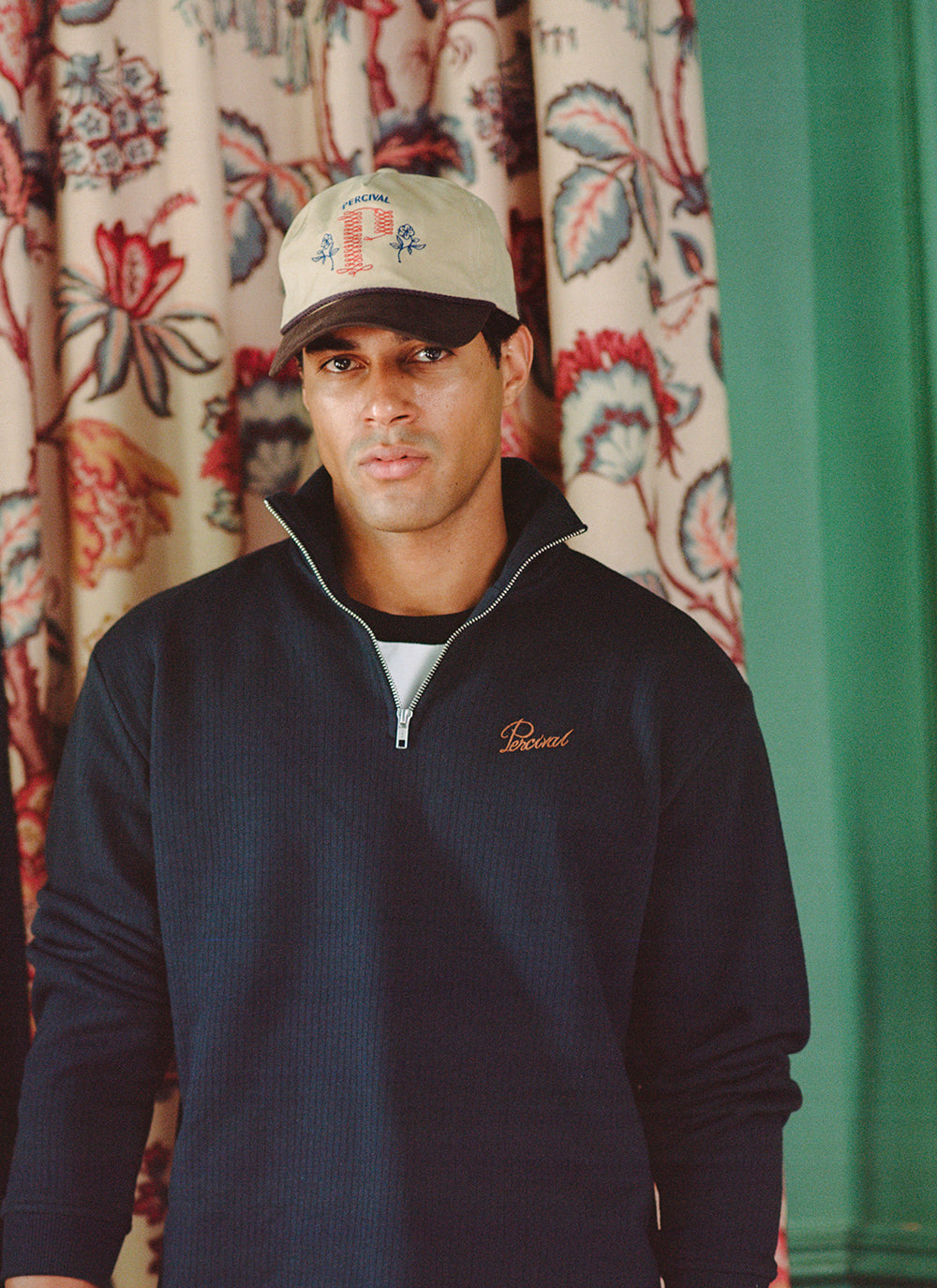 A model wearing a Percival navy cotton 1/4 zip jumper and a beige trucker cap against a patterned backdrop.